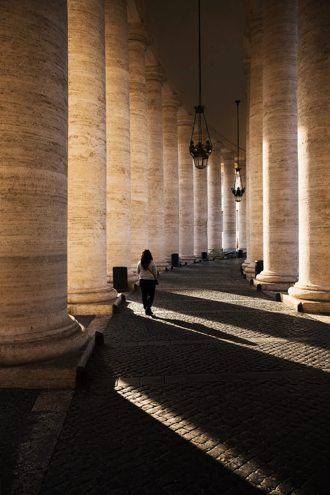 A woman walking through the colonnades of St. Peter's Square in Rome, with long column shadows on cobblestones